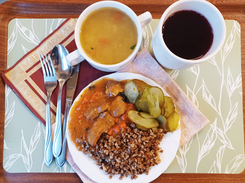 danie obiadowe na białym talerzu i na brązowej tacy, sztućce oraz kubek z zupą i kubek z napojem